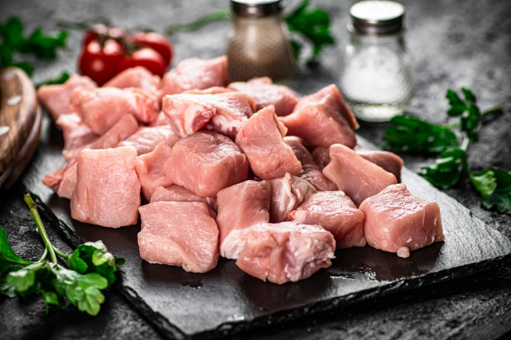 Pieces of raw pork on a stone board with parsley, tomatoes and spices.