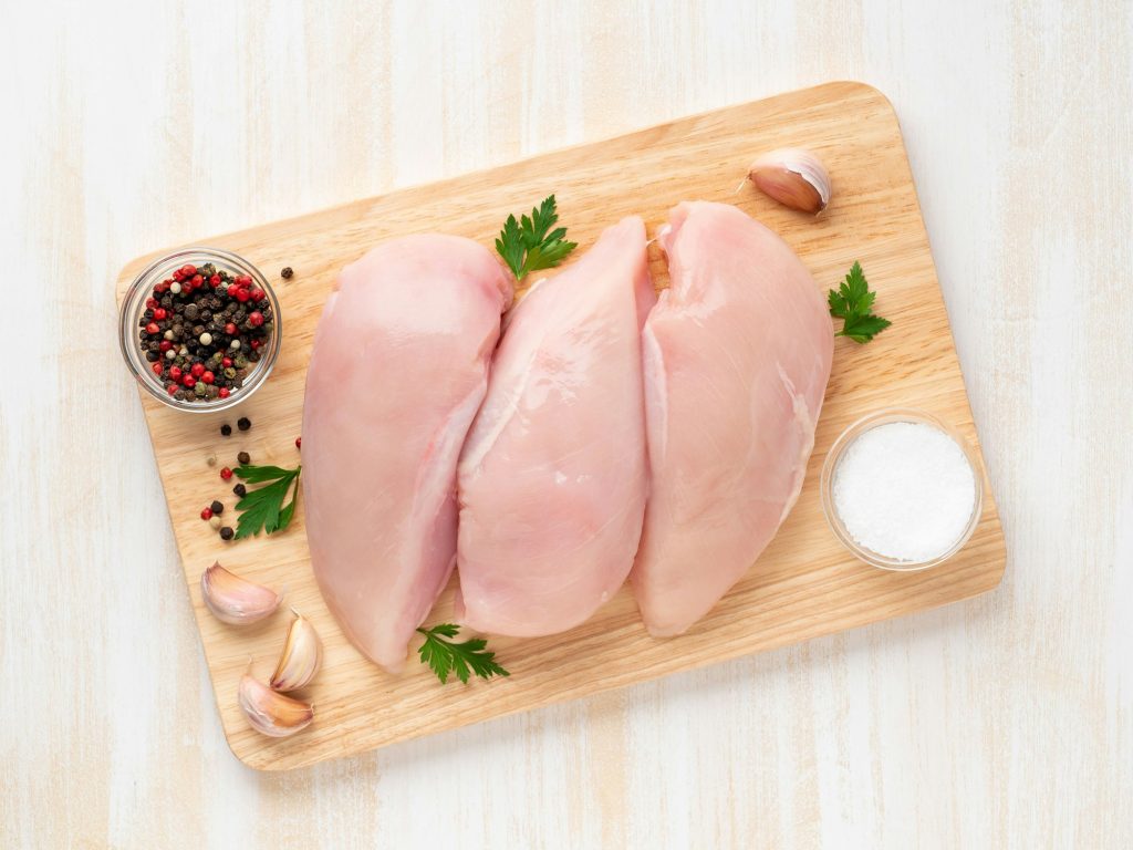 Top view of raw chicken breast fillet with spices on a wooden board on a white wooden table