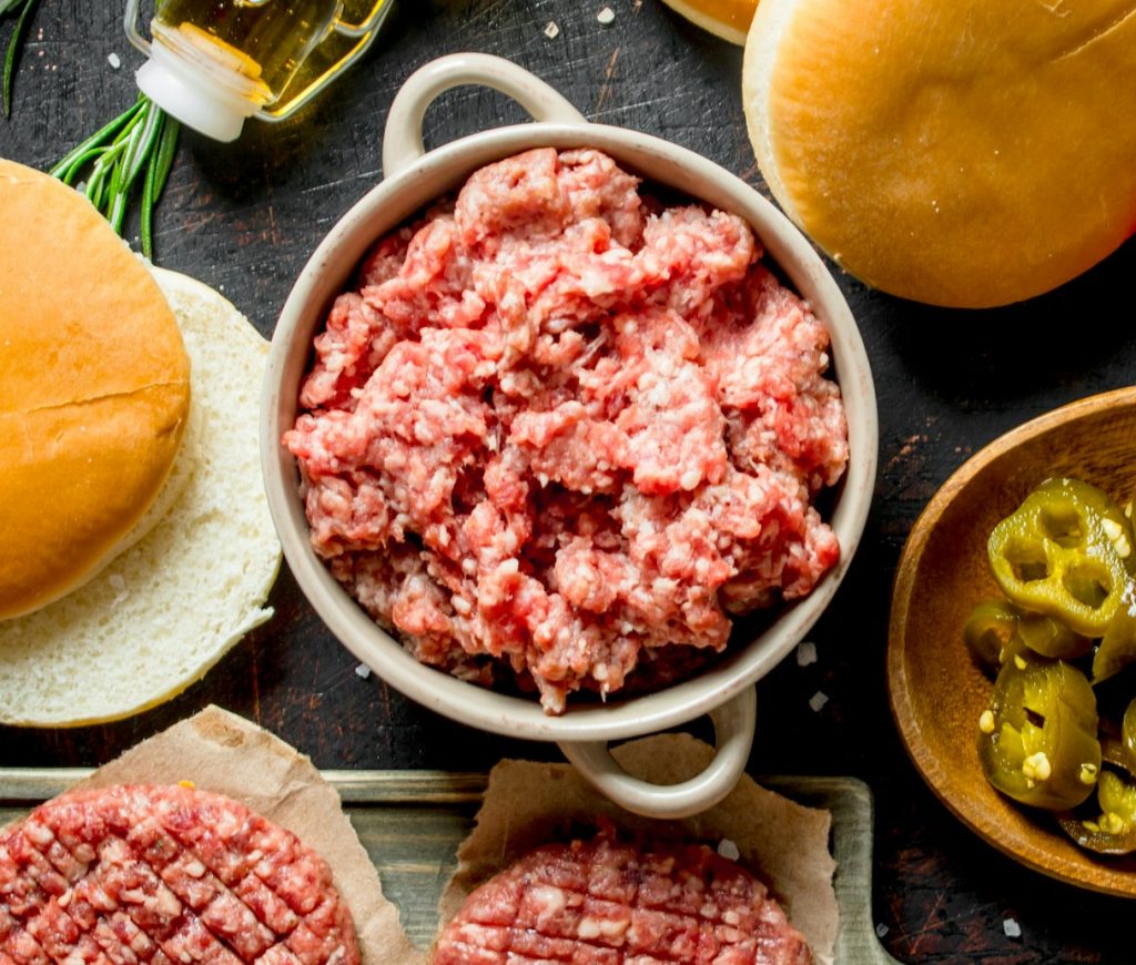Raw burgers with ground beef, oil, buns and tomatoes.